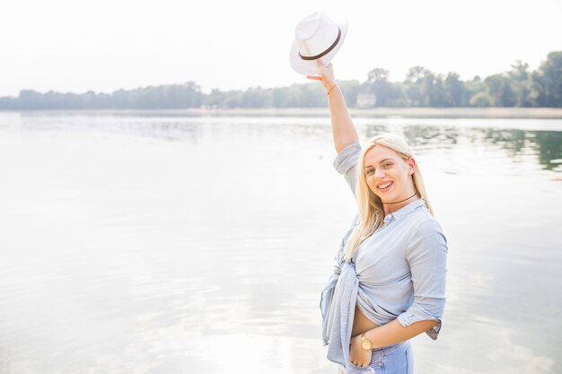 Smiling young woman raising hand holding hat standing near the lake