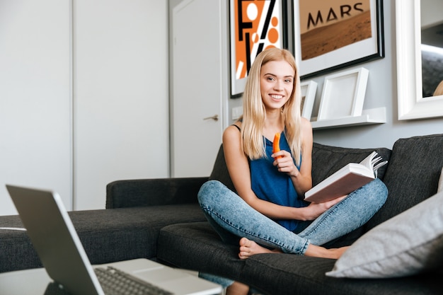 Free Photo smiling young woman reading book and eating carrots at home