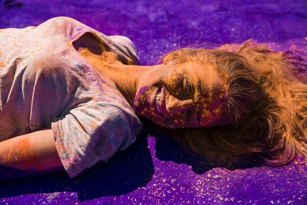 Smiling young woman's face covered with holi color lying on purple powder