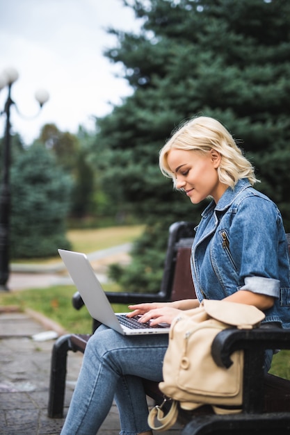 Free Photo smiling young woman sitting on the bench and use phone and laptop in the city autumn morning