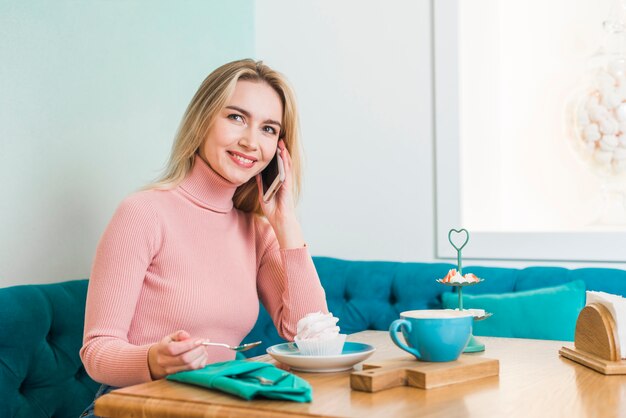 Smiling young woman sitting in cafe talking on mobile phone enjoying cupcake and coffee