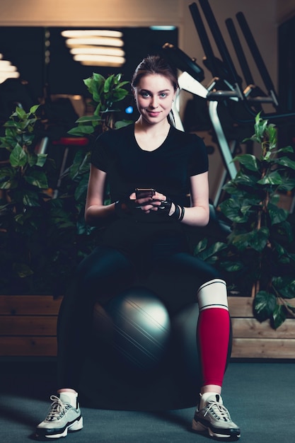 Smiling young woman sitting on exercise ball using cellphone