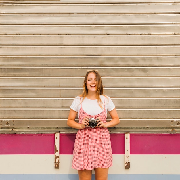 Smiling young woman standing in front of corrugated iron holding camera in hand