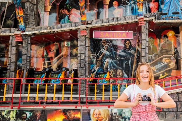 Free photo smiling young woman standing in front of haunted house fun ride holding camera