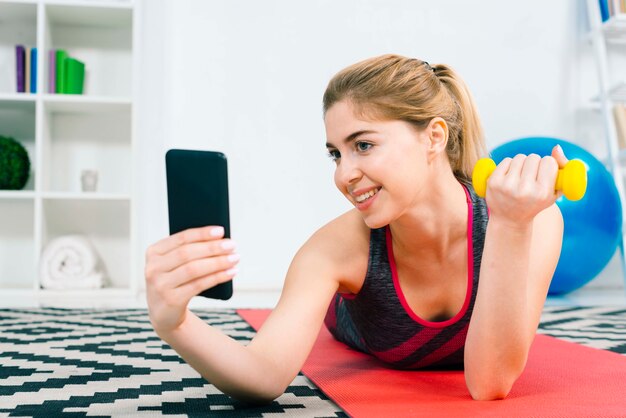 Smiling young woman taking selfie on mobile phone while doing exercise with yellow dumbbell