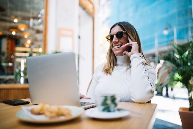 Smiling young woman using laptop at table with drink and croissants in street cafe