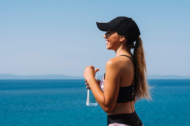 Smiling young woman wearing cap standing in front of sea holding water bottle in hand