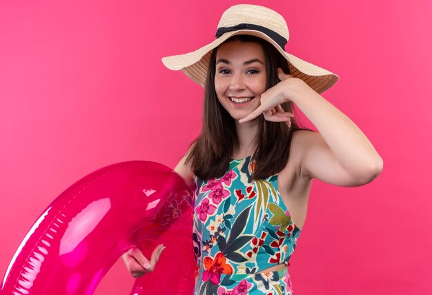 Smiling young woman wearing hat holding swim ring and doing phone sign on isolated pink wall