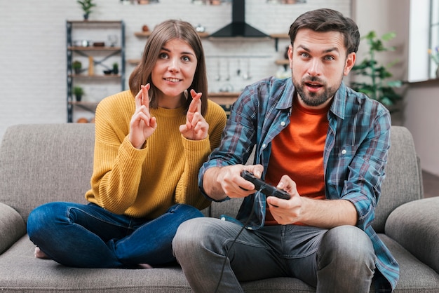 Smiling young woman with crossed fingers sitting near the man playing video game