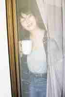 Free photo smiling young woman with cup of coffee looking through window