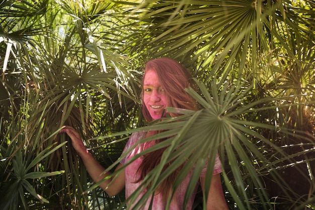 Free Photo smiling young woman with her face cover in holi color standing near the palm leaves