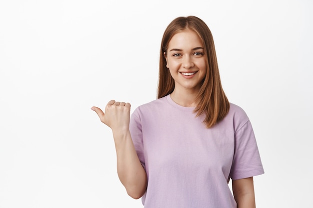 Smiling young woman, woman assistant pointing thumb left and look friendly, showing direction, logo banner on empty space aside, white wall