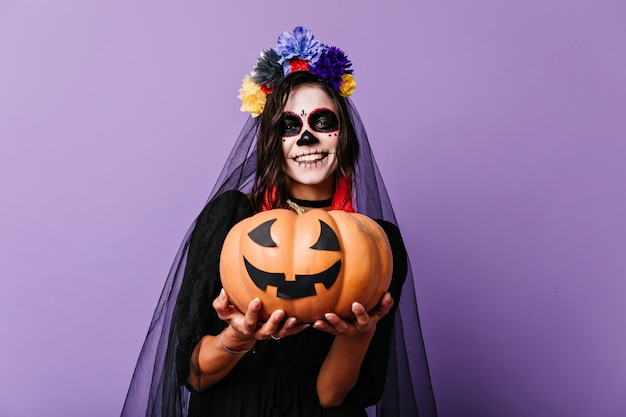 Smiling zombie girl in black veil posing on pastel wall. Glad woman in dead bride outfit holding halloween pumpkin.