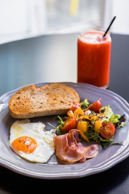 Free Photo smoothie in glass jar; toast; bacon; fried egg; salad on gray plate over the black table