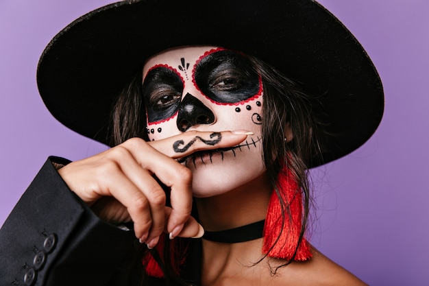 Snapshot of bright girl in wide-brimmed hat depicting Mexican man with mustache. Dark-haired lady posing on lilac wall.