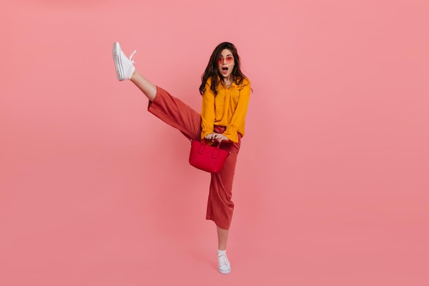 Snapshot of cheerful brunette in pink pants raising her leg high Lady in stylish outfit posing with handbag on isolated background