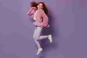 Free photo snapshot in full growth of fun girl jumping with longboard. curly woman posing in white jeans and pink coat.