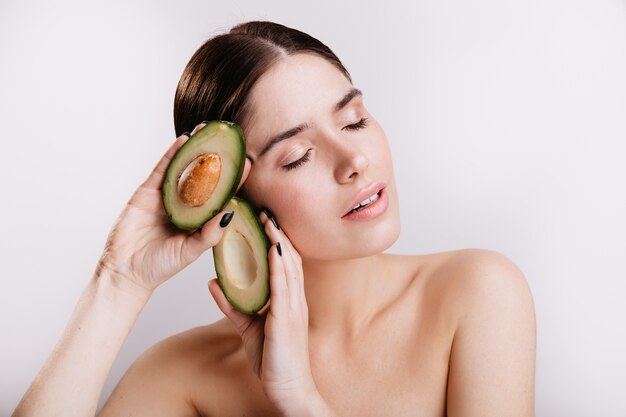 Snapshot of girl without makeup on white wall. Lady with closed eyes keeps healthy avocado.