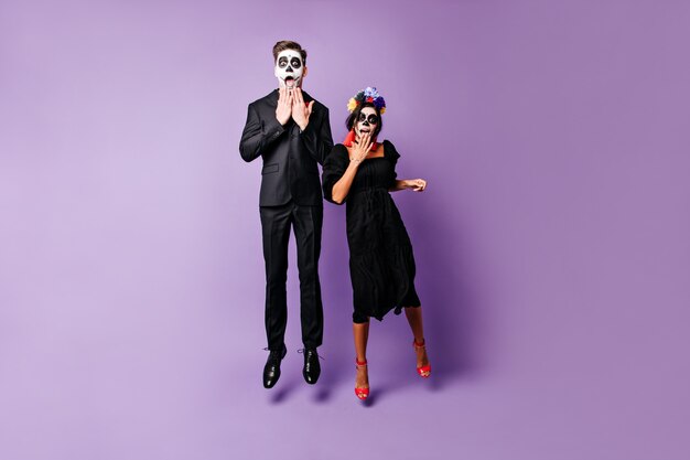 Snapshot of young slender guy and girl with painted faces in black outfits. Couple from Mexico looking surprised into camera, jumping on isolated background.