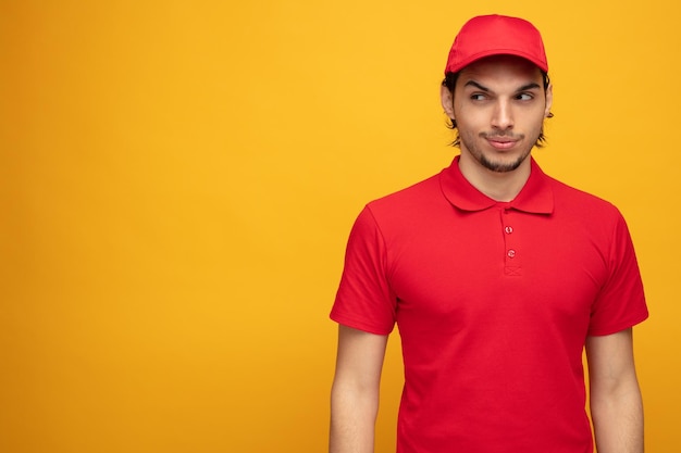 Free photo sneaky young delivery man wearing uniform and cap looking at side having some evil plans isolated on yellow background with copy space