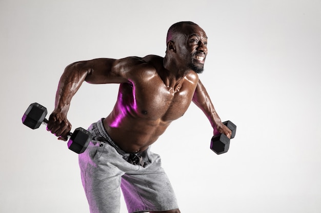 So growing the wings. Young african-american bodybuilder training over grey background. Muscular single male model in sportwear with weights. Concept of sport, bodybuilding, healthy lifestyle.