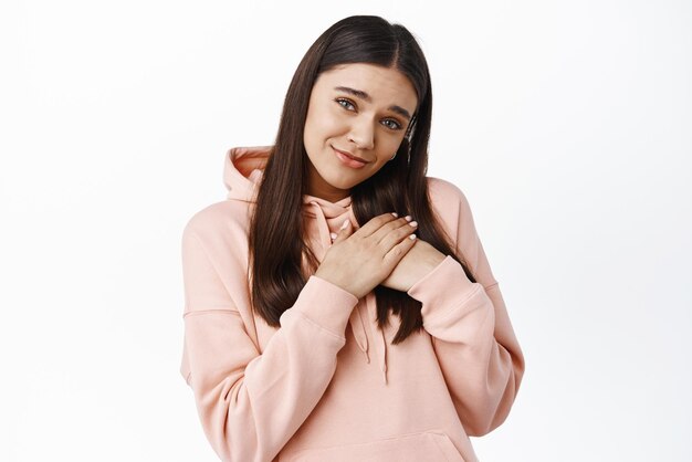 So sweet and cute Heartfelt young girl melting from cuteness holding hands on heart and smiling at camera with gentle and romantic expression white background