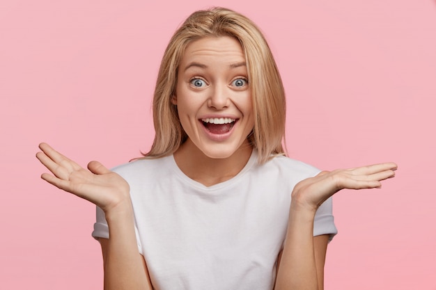 So, what? Happy woman shrugs shoulders, looks joyfully with her blue eyes, dressed casually, isolated over pink wall. Hesitant cheerful blonde female expresses positive emotions and feelings