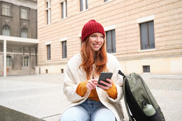 Free photo social media and people young redhead girl sits on street uses mobile phone app looks up information