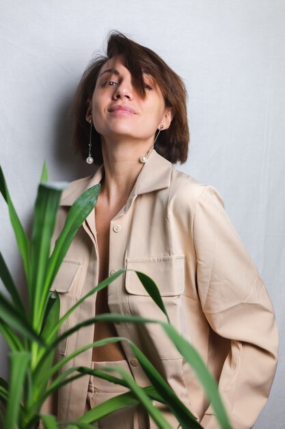 Soft indoor portrait of caucasian gentle woman wearing beige suit no bra, posing behind palm tropical plant,  gray.