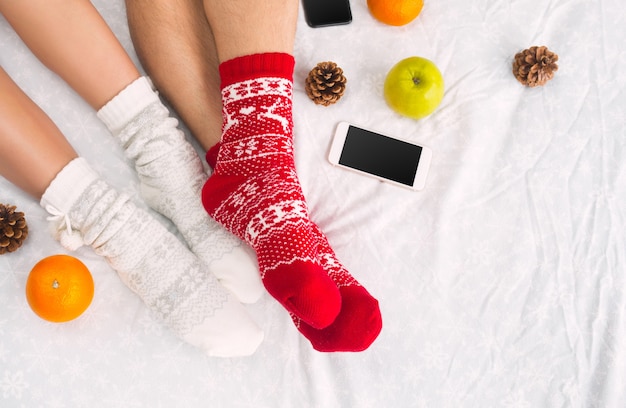 Free photo soft photo of woman and man on the bed with phone and fruits, top view point. female and male legs of couple in warm woolen socks.