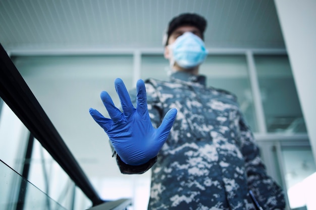 Free photo soldier in camouflage uniform wearing protective gloves and mask showing stop sign at hospital entrance