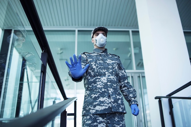 Free photo soldier in military uniform with rubber gloves and face protection mask guarding hospital and gesturing stop sign