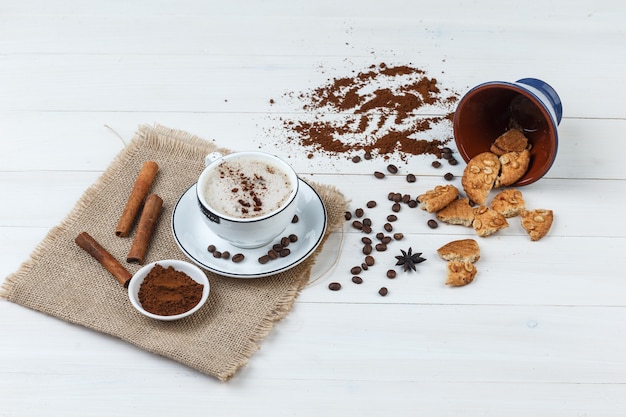 Some coffee with coffee beans, grinded coffee, cookies, cinnamon sticks in a cup on wooden and piece of sack background, high angle view.