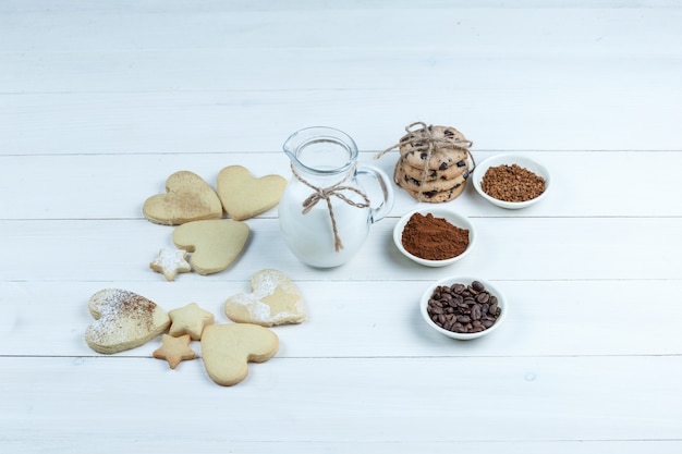 Some different types of cookies with coffee beans, instant coffee, cacao, jug of milk on white wooden board background, high angle view.