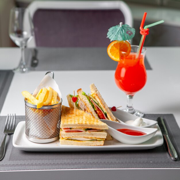 Some fast food with sandwich, french fries, red cocktail, fork and knife on table, side view.