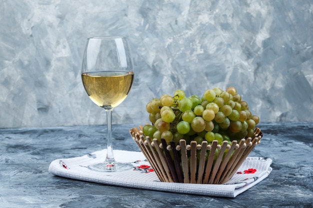 Some green grapes with glass of wine in a basket on grungy plaster and kitchen towel background, side view.