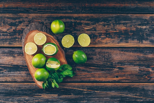 Free photo some green lemons with slices and leaves on wood slice and dark wooden background, flat lay. space for text