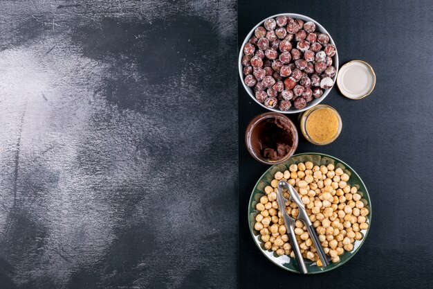 Some of shelled and cleaned hazelnuts with cocoa spread and nutcracker in a white bowl on dark stone table, top view.