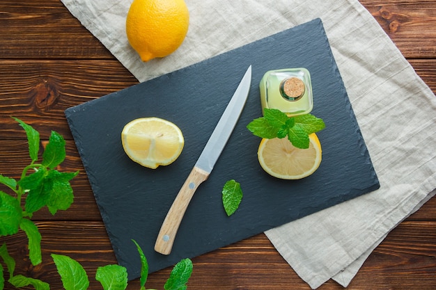 Some sliced lemon with black cardboard, juice bottle, wooden knife in a bowl on wooden surface, top view.