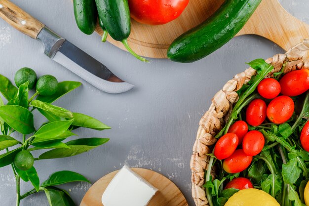 Some tomatoes with knife, lemon, cucumber, cheese, greens in a cutting board on gray surface, top view