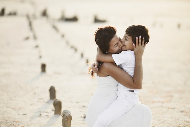 Son is biting mother's nose and hugging her, dressed in white clothes, sitting on the sand