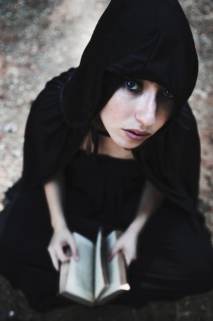 Free photo sorceress in black hood with opened book sitting on ground