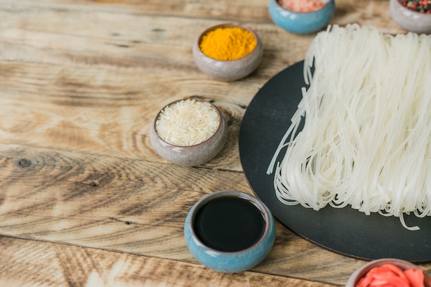 Free photo soya sauce; raw rice; turmeric in bowl near dried rice noodles on black tray over wooden texture background