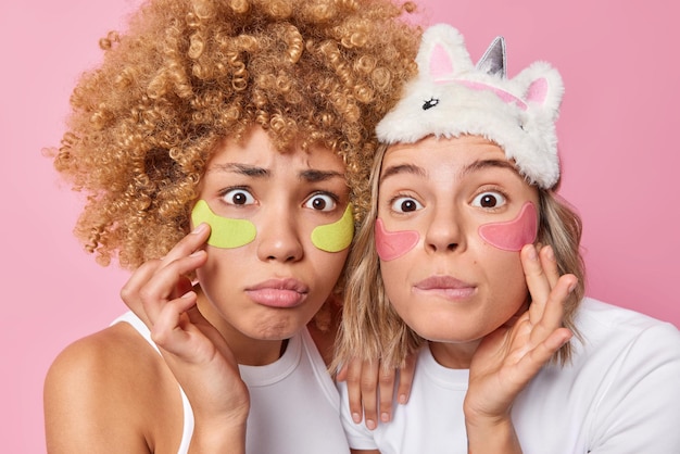 Free photo spa day shocked young friendly women apply beauty patches under eyes for anti aging touch face gently look wondered at camera dressed in casual domestic clothes isolated over pink background