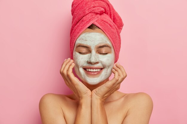 Spa girl applies clay mask on face, keeps eyes closed, touches cheeks, gets pleasure from beauty procedure, refreshes skin, smiles positively isolated on pink wall. Relaxation, healthy lifestyle