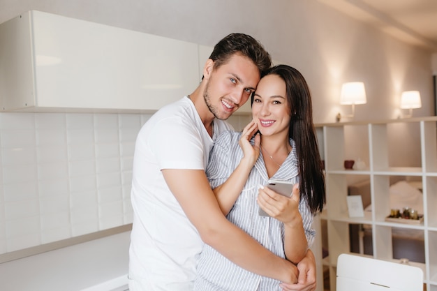 Spectacular brunette woman with cell in hand posing in husband's arms with smile