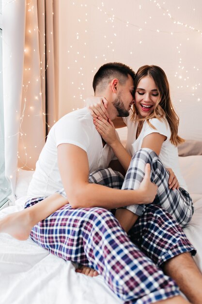 Spectacular girl in checkered pants playfully posing in bed with husband. Good-humoured guy embracing his wife in morning.