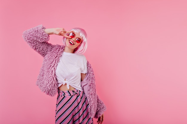 Free photo spectacular girl in shiny pink peruke laughing during indoor photoshoot. charming young woman in bright fluffy jacket expressing positive emotions