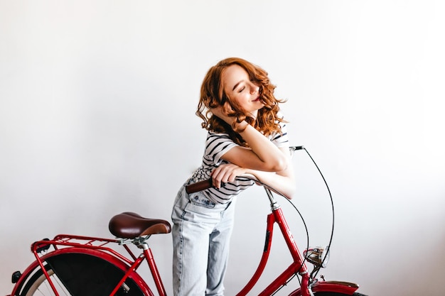 Free photo spectacular girl in striped tshirt posing on white background with eyes closed inspired ginger woman standing near her bicycle