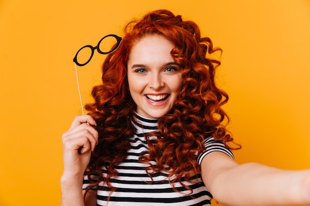 Free photo spectacular redhaired girl in great mood makes selfie and smiles portrait of blueeyed woman in striped top with toy glasses